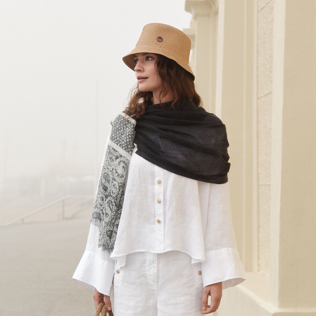 Busnel ss25 model image standing in misty coastline wearing white linen clothing with marine printed scarf and natural colored straw hat