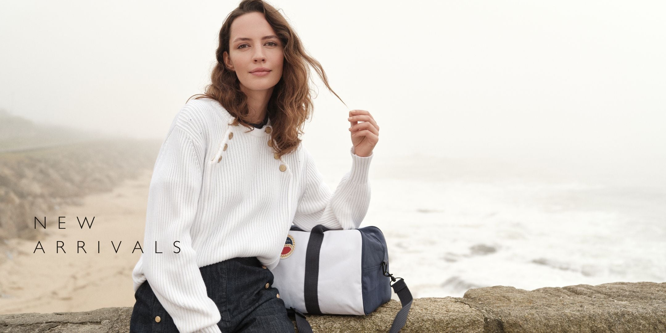 SS25 campaign image. Model sitting with coastline backdrop, wearing knitted sweater