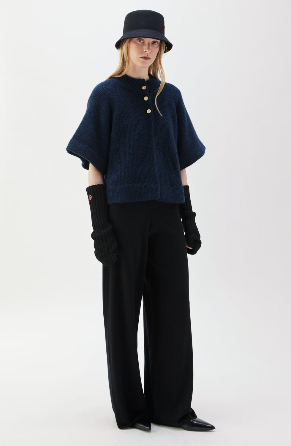 A model wearing a dark blue melange cape-styled cardigan with a round neck, short flutter sleeves, and golden buttons. Also wearing black pants and a matching hat.